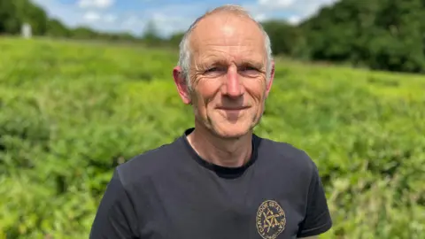 A man stands in a field of tea plants on a sunny day. He is wearing a dark T shirt and the plants are behind him.