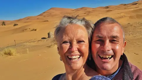 Karen Davies Image of a tanned couple who are hugging and smiling at the camera. In the background can be seen desert sand dunes.