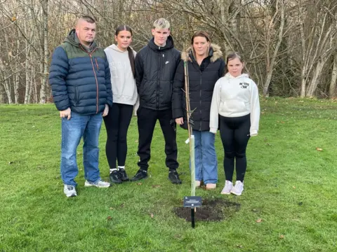 BBC/Alison Freeman Holly's family stand next to a thin sapling that has been planted in the ground. There are two adults - Micala, who has blonde and brown hair and is wearing a thick coat and jeans, and Lee, who is wearing jeans and a puffer jacket and has a short stubble beard and short shaved hair - and three children - an older teenage boy and girl and a younger girl.