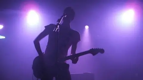 Andrew Woodger/BBC Snow Patrol lead singer Gary Lightbody on stage at Latitude Festival in 2006. He is singing while playing the guitar. He is silhouetted as purple lights shine from behind.