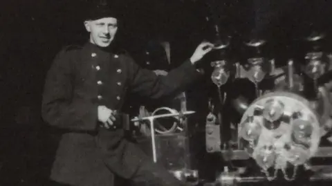 William Cruickshank A black and white picture of William Crockett standing on a piece of machinery. He is wearing a dark jacket with buttons on either side.