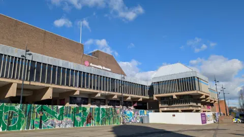 Large grey brick building with long rectangular windows and a wall infront of it with graffiti 