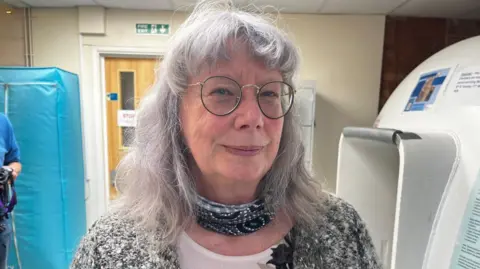 A woman with long grey hair and glasses is looking into the camera. She is wearing a grey cardigan with a large flower brooch. 