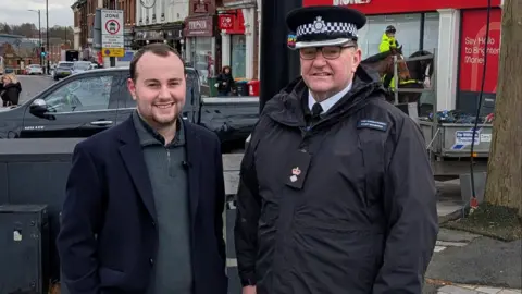 A man in a blue jumper and blazer next to a police officer in a town centre - it is Councillor Wilton and Ch Sup Bainbridge