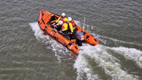 LDRS An orange in-shore lifeboat and crew wearing high vis, cutting through calm water leaving a foamy trail
