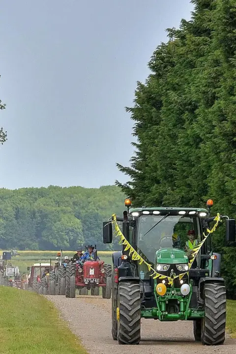 Derek Bell Photography A green tractor is decorated with yellow bunting. It has a trail of tractors behind it and is on a tarmac road with grass and trees to the side. There are also trees in the background.