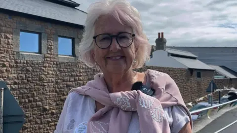 A lady with grey hair and black glasses smiles at the camera. She wears a white short sleeve top with a pink jumper with white embroidered details on it draped over her shoulder. In the background home-made bunting attached to railing blows in the wind.