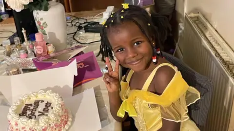 A smiling Mayar Yahia wears a yellow dress and beaded plaits. She is sitting in front of a cake which says Happy Birthday Mayar