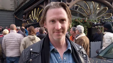 A man with long fair hair wearing a black leather jacket and denim shirt. He has stubble. Behind him is a bronze statue of the musician Rory Gallagher. There are people gathered around looking at it and taking pictures. 