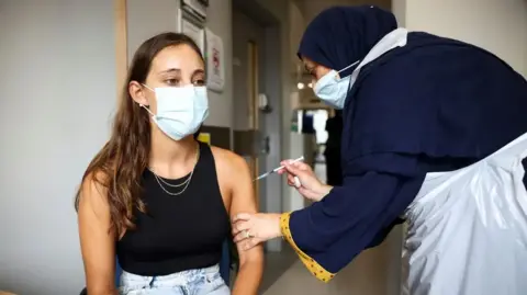 A woman is sat down being injected. She is wearing a black vest top and a mask and jeans. The nurse, also in a mask, is bending over holding her left arm.