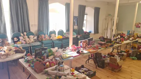 Chris Ellis Tables filled with toys and teddies at the church. There are teddies displayed on stacked blue chairs. The floor is brown laminate and there are three large windows with curtains behind the chairs