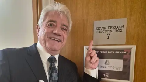 A man with white hair and a black blazer, black and white spotty tie and white shirt (Kevin Keegan) is smiling and pointing to a door which says Kevin Keegan, executive box 7 on it.