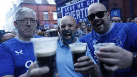 Reuters Three male Everton fans dressed in blue hold plastic pints of Guinness aloft