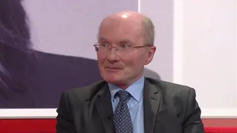 Professor John Bryson - a clean-shaven balding man with glasses is weaing a dark grey suit with a blue shirt and tie and sat on a red Midlands Today sofa and looking to the left