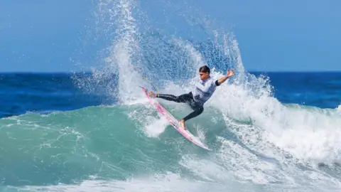 A person on a pink surfboard is carving sharply across a wave, sending a large spray of water into the air. The ocean stretches into the background under a clear blue sky, with the surfer positioned mid‑turn on a breaking wave.