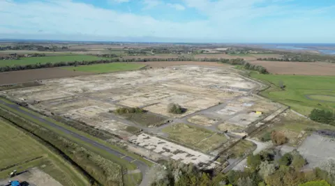 A drone view of a disused gas terminal. Little remains except roadways and hard standing. The extensive site is bordered by farmers' fields. In the distance, hedgerows and trees give way to a beach and the blue sea.