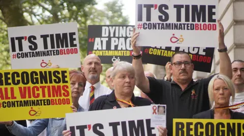 PA Media File photo dated 26/07/23 of campaigners, including many who are personally infected and affected by infected blood, gathering in Westminster, London. The group, posing for the camera, hold white and yellow signs which read 'It's time', 'Dying at pace' and 'Recognise all victims'.