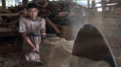 Getty Images A large spinning blade cuts through a log of wood with a worker, covered in sawdust, controlling the process at the back of the shot. 
