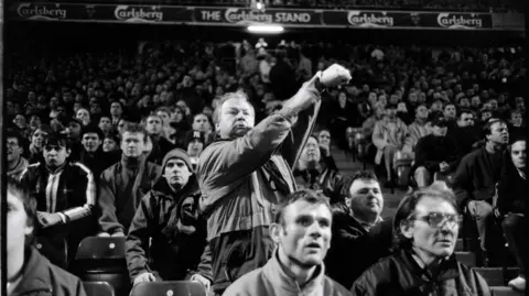 A close-up, shot in black-and white, of a football fan standing up and pointing to his watch while other supporters around him remain seated. 