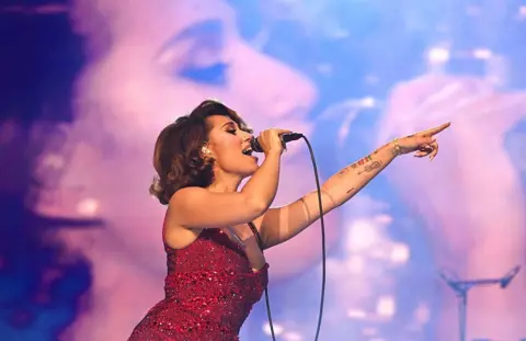Getty Images Raye performs on stage during her 'This Tour May Contain New Music' tour at The O2 Arena on May 19, 2026 in London, England. She is wearing a red, sparkly gown and is singing into a wired microphone. The backdrop is an enlarged video screen