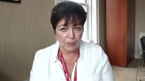 Rita has short brown hair and blue eyes. She is wearing a white blazer and red lanyard. She is looking at the camera. Behind her is a chest of drawers and a chair.