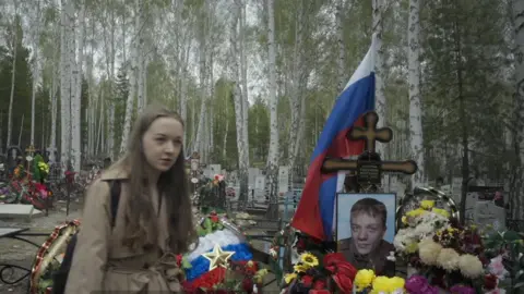 Pavel Talankin/BBC Storyville/Made in Copenhagen young girl with long brown hair in beige mac at graveside of her brother, which has a photo of him, the Cross, Russian flag, surrounded by yellow, white red and dark pink flowers. Tall trees in the background