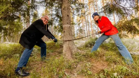 The two mayors stand on either side of a tree trunk with a two-man saw. They look at the camera and smile with their legs braced. Mayor Anne Lindboe wears a hat and orange coat and Mayor Paul Dimoldenberg wears a black coat with blue jeans.