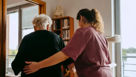 An aged female pinch short achromatic hairsbreadth is escorted by a female pinch agelong brown, wearing maroon scrubs. The younger of nan 2 has her near manus connected nan woman's back. The aged female wears a achromatic dress. A woody book shelf, 2 shelves wide and 4 agelong is partially stacked pinch books. There are 2 windows either broadside of nan bookshelf, a assemblage of h2o is visible outside.