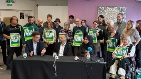 A group of people at a table speaking into mics while people behind them hold signs which say "Green Party".
