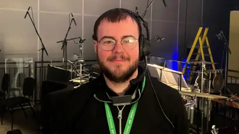 Photograph of Reuben Rowlands, who is working on Angel's Bone. He is pictured in front of musical instruments at the Aviva Studios.