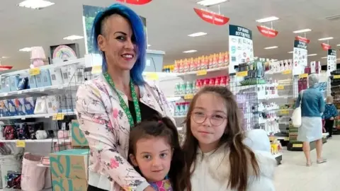 Clare Smyth Kaleea, who has long brown hair and pink-rimmed glasses, is wearing a white top and has her arm around her younger sister, Dakota. Dakota, who also has long brown hair, is dressed in a pink and purple top and smiling. Clare Smyth, who has long blue hair, is wearing a lanyard, a multicoloured jacket and a black top. She is standing inside a shop.