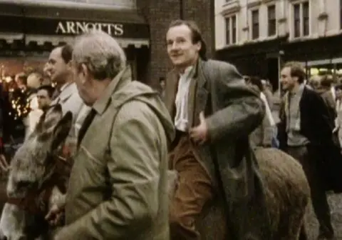 David Hayman in a long coat is on a donkey in Sauchiehall Street. The shop Arnotts is behind him.