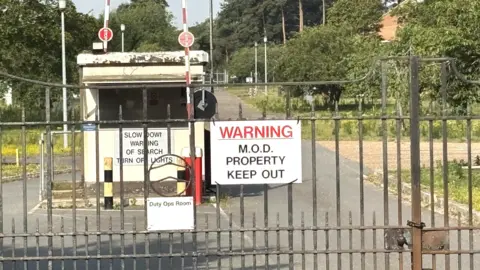 A gated entrance to a Ministry of Defence site, with raised barrier arms and signs reading “WARNING M.O.D. PROPERTY KEEP OUT” and “Slow Down! Warning of Search Turn Off Lights,” stands amid trees and roadside vegetation.