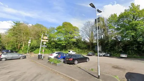 Car park in Wetherby showing about 10 parked cars, with trees and floodlights.