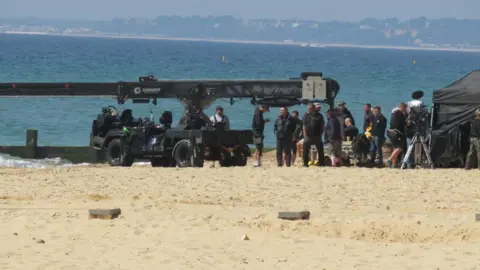 Newsquest Film crew on a sandy beach beside the sea, with a large camera crane, production equipment, and crew members preparing for a shoot.