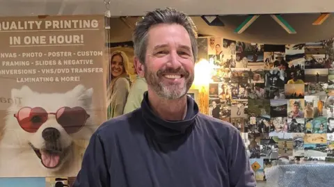 Simon Stockdale smiles as he stands in front of the glass window of his print shop. He has grey-brown swept back hair and a beard.