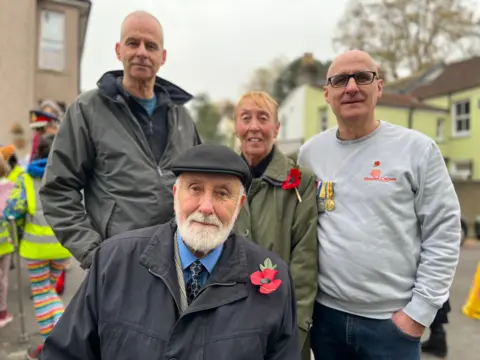 Four members of LCpl Room's family. A tall man in a grey jacket, a woman with red/blonde hair in a green coat with a poppy, a man in a grey sweatshirt with some medals pinned to it and an older man with a beard in a shirt and tie and coat, with a poppy pinned to it.