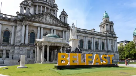 Getty Images Belfast City Hall is a large white building with pillars and designs. There's a large statue of Queen Victoria at the front. Belfast in large letters are displayed on grass at the front of the building.