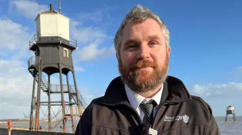 Stuart Woodward/BBC Will Lodge is smiling and has grey hair and a beard. He is wearing a black zip-up jacket over a white shirt and black tie with white polka dots. The lighthouses are pictured behind him.
