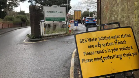 Jack Fiehn/BBC A yellow sign by SES water explaining the bottled water one way system sits by a wall, the entrance to Bourne Hall car park can be seen. In the distance, cars are queuing up for bottled water.