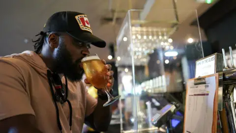 Getty Images A man drinks a pint at a pub in Newcastle upon Tyne, England. He is wearing a baseball cap and a hooded t-shirt.