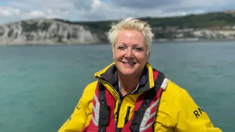 Simon Jones/BBC Paula Lain - A woman with short blond hair in yellow RNLI seeds and a red survivor jacket. The photo was taken from a boat in the sea. In the background is the white slope of Dover.
