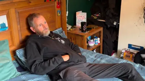 LDRS A bearded man wearing a black hooded top and grey jeans lying on a bed with his hands inside his pockets. There is a black water cup on a brown wooden bedside table. 