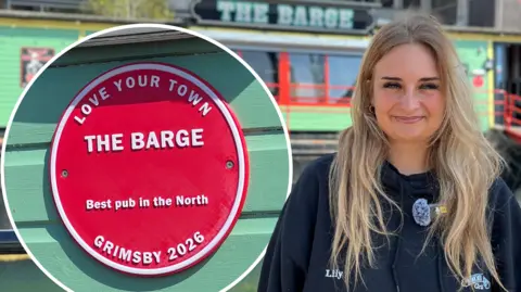 Woman with long blonde hair smiles to camera. She is wearing a dark, navy jacket with her name 'Lily' embroidered into it. In the background The Barge, a bright mint green boat with red detailing around the glass windows can be seen. There is also a close-up picture of the red plaque which reads: 'Love Your Town, The Barge, Best pub in the North, Grimsby 2026'.
