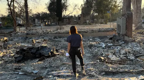 Harry Butler A woman standing in the middle of debris from the wildfires. In the background are trees and a fence. To the right is what looks like the bricks from a fireplace.