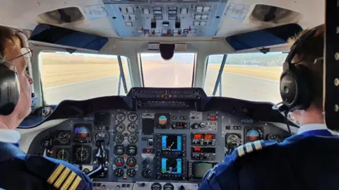 Two pilots sitting in a plane cockpit. They are staring out of the front window, facing a runway. The pair are surrounded by switches, dials and steering equipment.