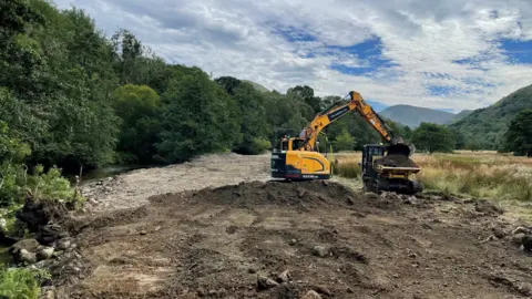 Tom Lindsay A digger on the banks of the river, removing earth and placing it on the back of a dump truck.