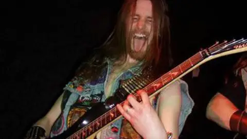 Darren Parkinson A man with long brown hair poses with an electric guitar. He has his tongue hanging out and his eyes screwed up. He is wearing a denim vest with colourful badges