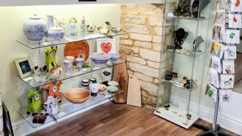 Tarot Crick Interior of an art gallery with paintings on pale walls, glass display cases holding ceramics and jewellery, wooden shelves with bowls and decorative pieces.