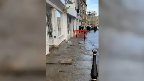 Water flowing down towards Market Square in Durham city centre. An orange barrier has been erected and there are people gathered on the pavement.